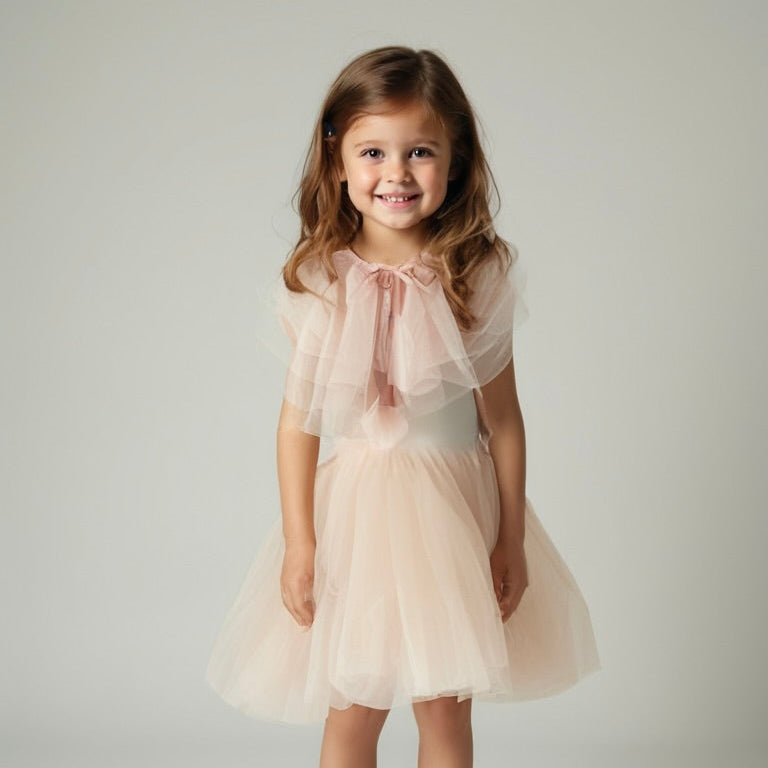Young girl wearing a light pink dress with a sheer overlay against a plain background