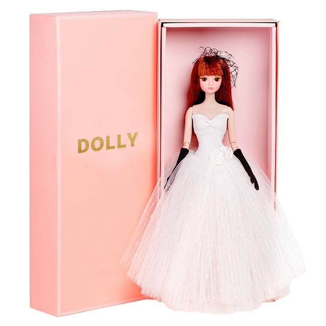 A DOLLY® bride fashion doll with white wedding tutu dress, straight red hair, pearl hairpiece, and long black gloves, presented in a pink DOLLY® giftbox.