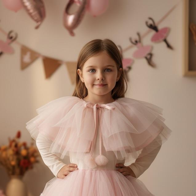Young girl in a pink dress with ruffled sleeves and cape, standing in front of decorative balloons and flags.