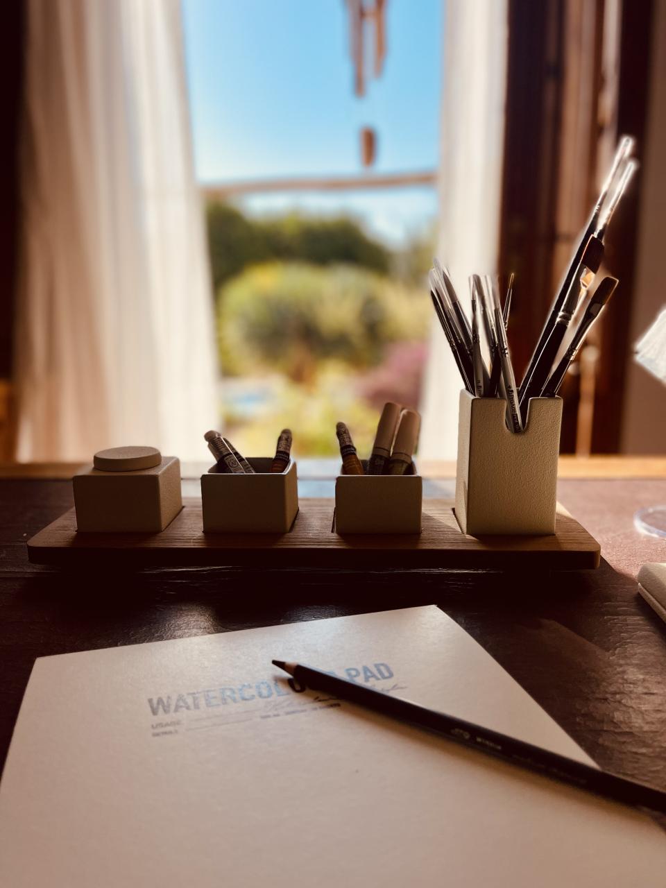 Desk setup with stationery items and a watercolor pad in front of a window with a view.