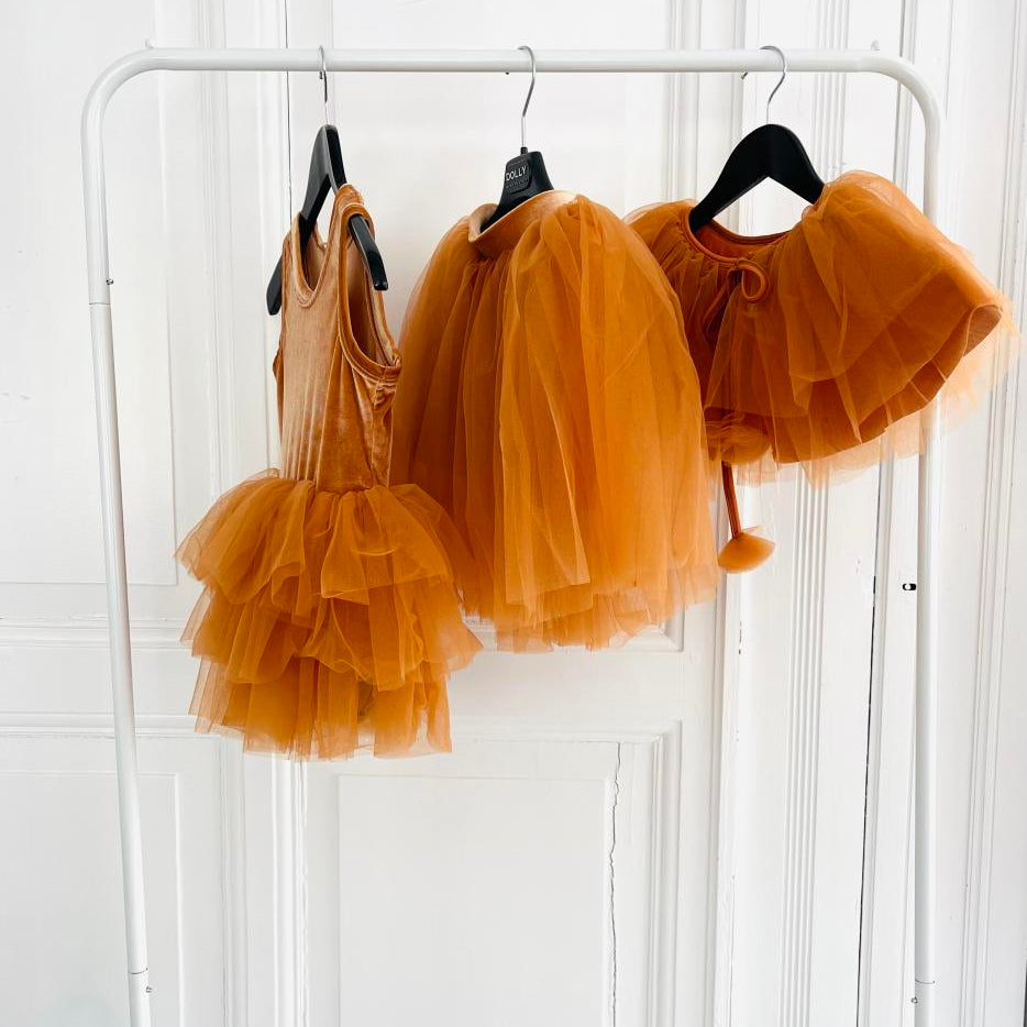 Three orange children's dresses with layered skirts hanging on a rack against a white background.