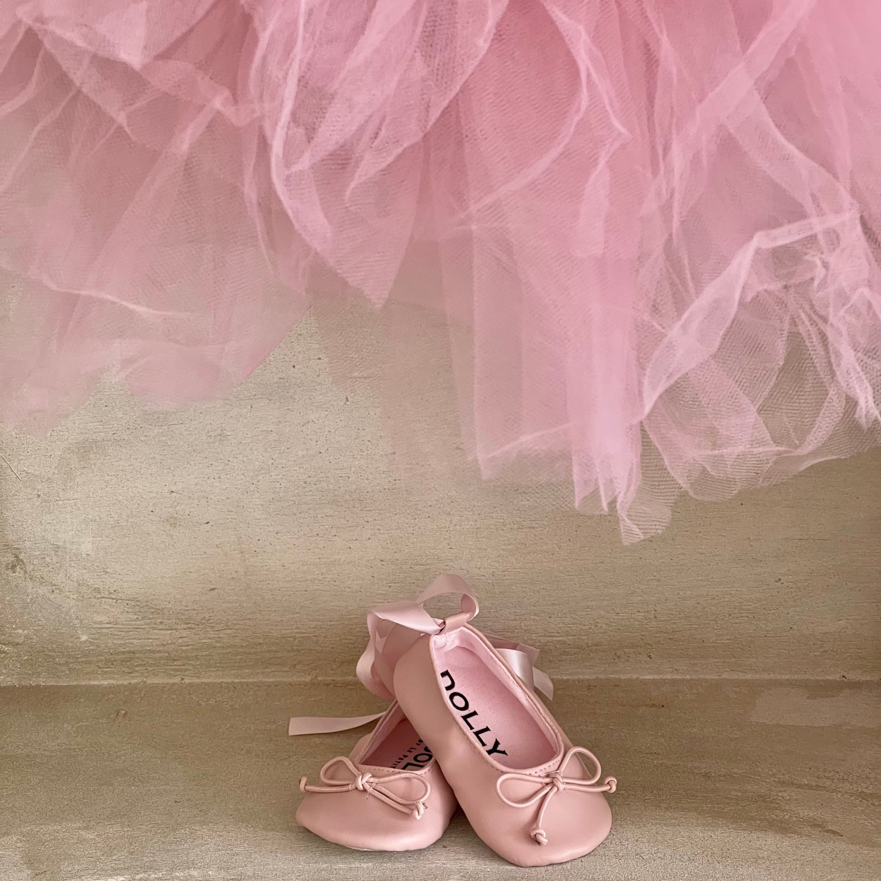 Pink ballet shoes with ribbons on a wooden floor, partially covered by a pink tulle skirt.