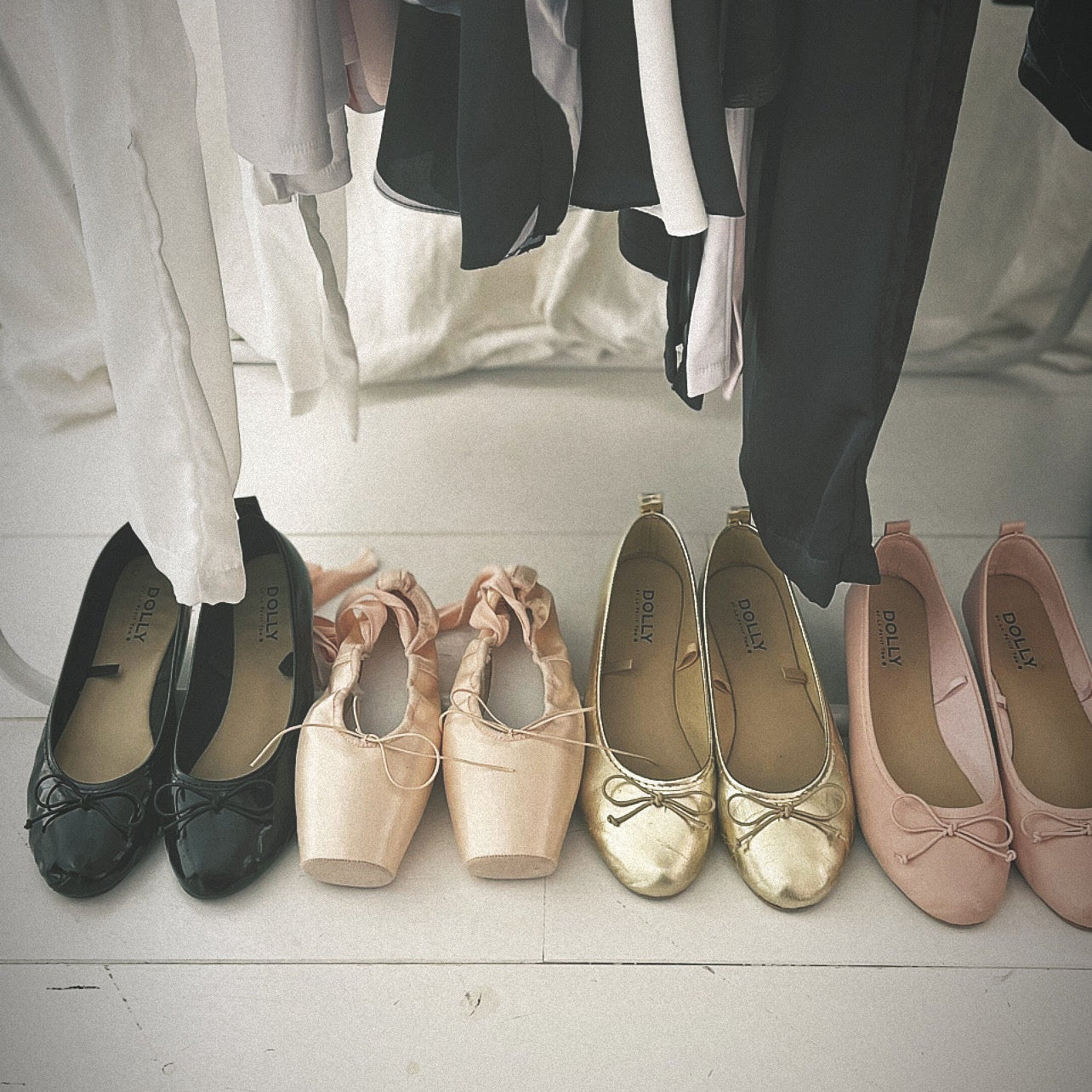 Set of flats in black, beige, gold, and pink on a light wooden floor with hanging clothes in the background.