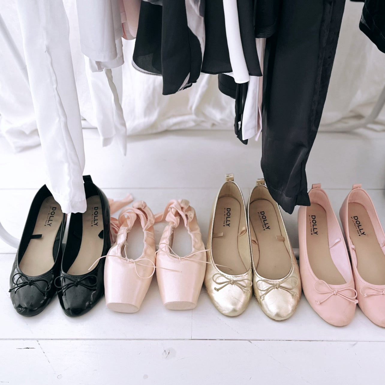 Set of black, pink, and gold flats on a white floor with hanging clothes in the background.