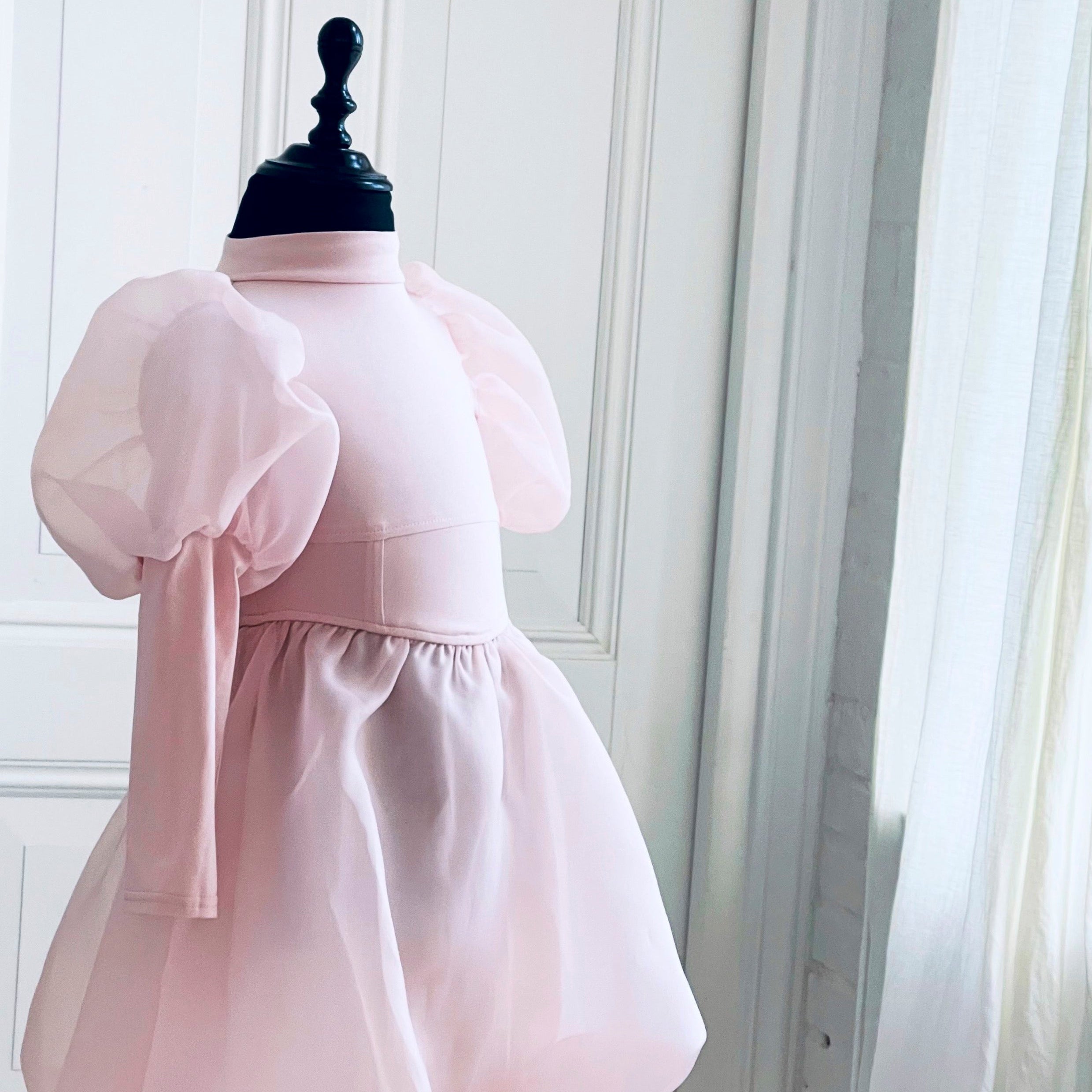 Pink dress on a mannequin against a white wall with a curtain.