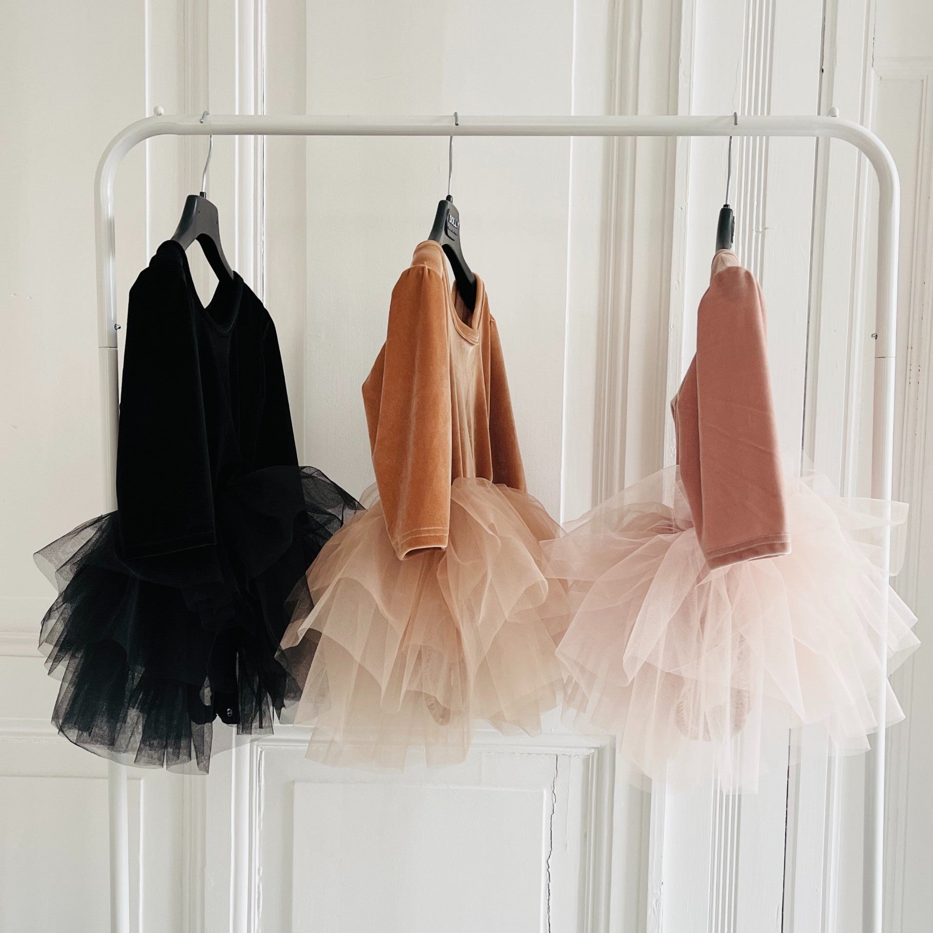 Three children's dresses with tulle skirts hanging on a rack against a white wall.
