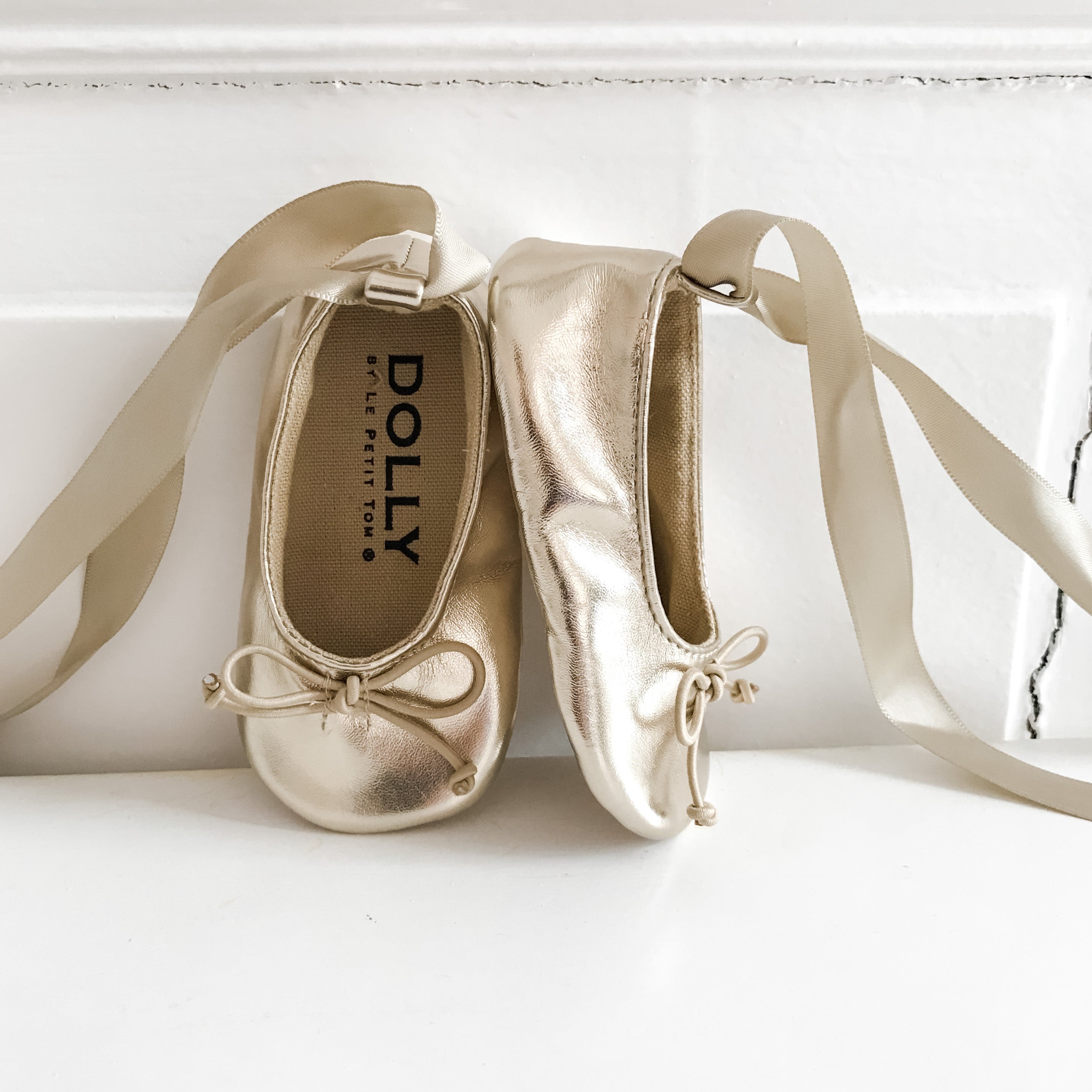 Pair of gold children's shoes with ribbons on a white surface, branded 'Dolly'.