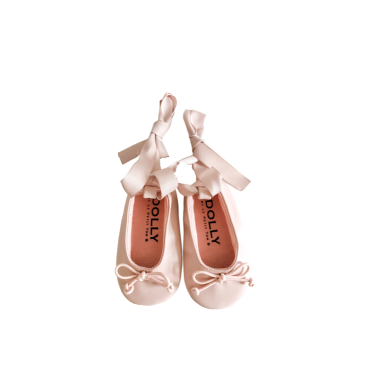 A pair of pink DOLLY by Le Petit Tom baby ballerina shoes with ribbons tied in bows, displayed against a white background with the brand name 'DOLLY' visible on the insole.