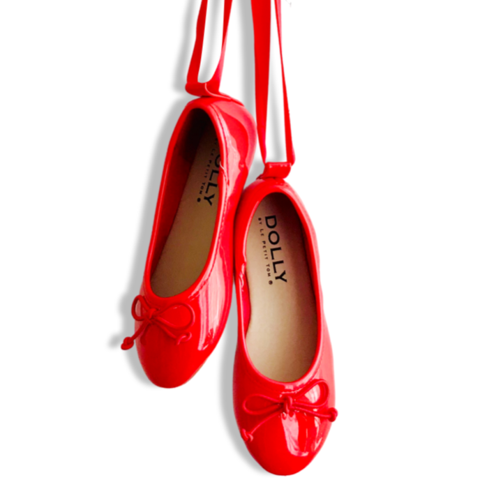 A pair of red DOLLY by Le Petit Tom ballerina flats with satin ribbons tied in bows, hanging against a white background.