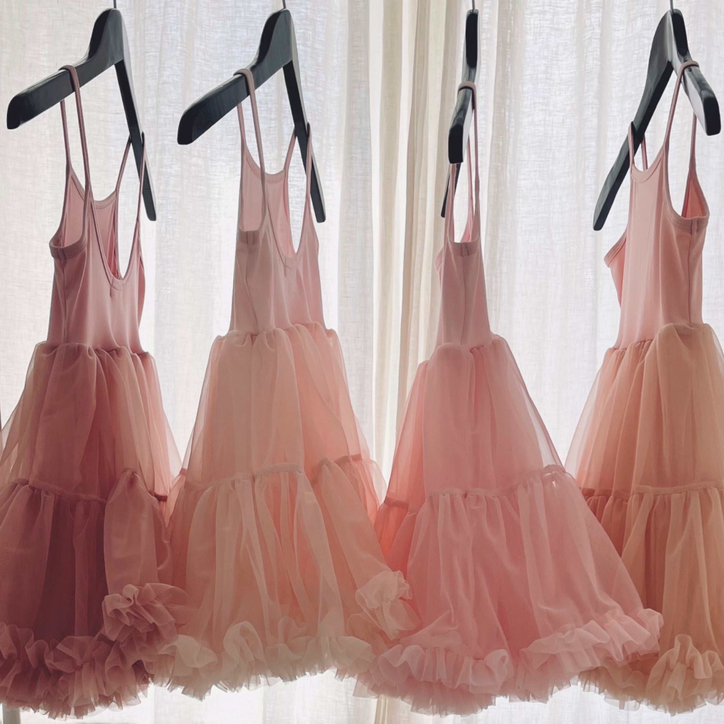 Three pink tiered dresses on hangers against a white curtain background